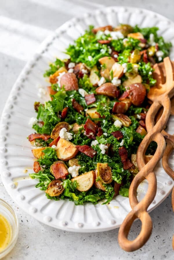 wooden salad tongs next to a potato brussels sprouts salad on a large white platter.